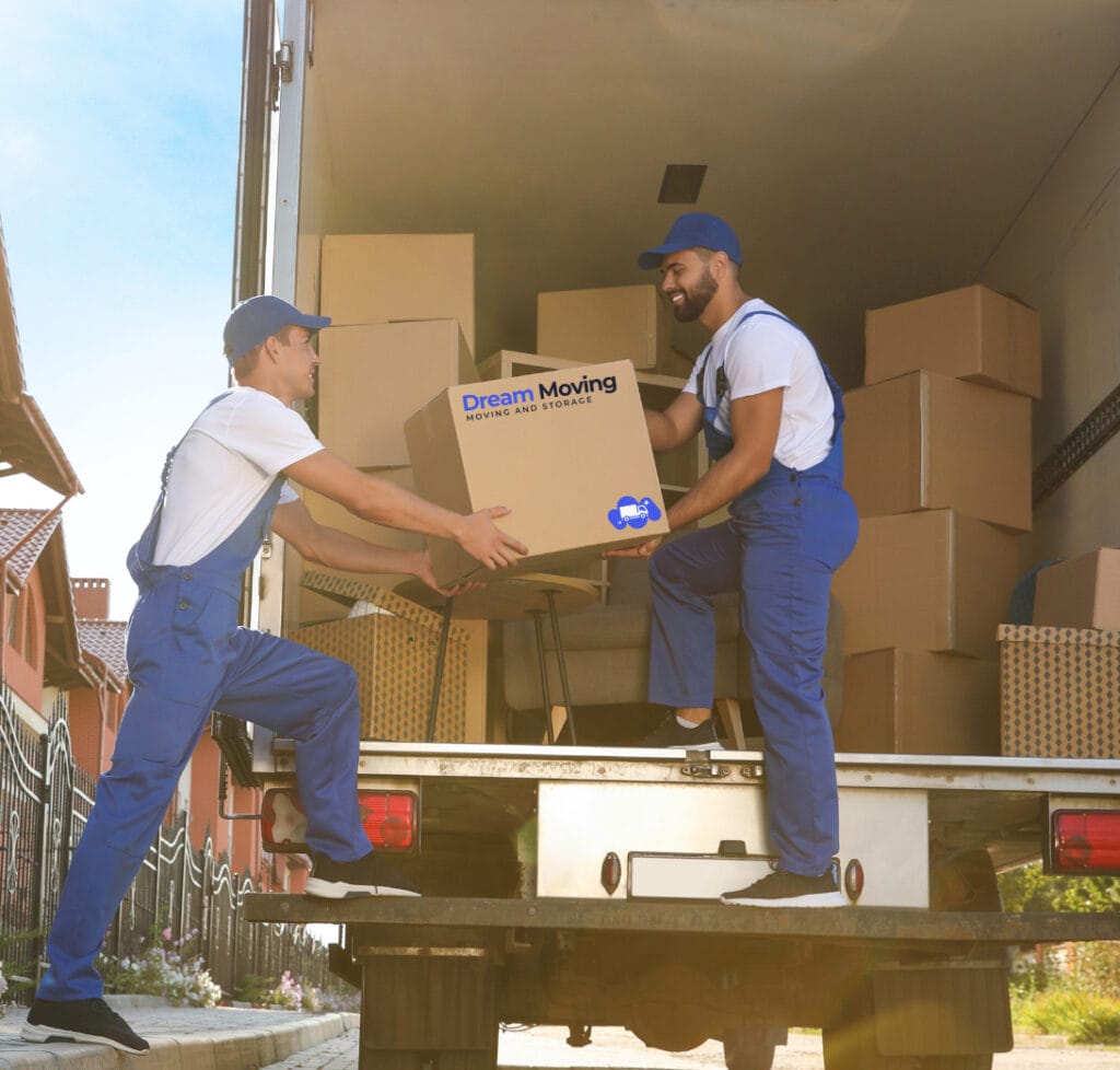 NYC office moving company. Two Dream Moving members wearing blue uniforms load cardboard boxes into a moving truck parked in a residential neighborhood. One person stands inside the truck, reaching down to receive a box from the other standing on the pavement. The box displays the text “Dream Moving” and “MOVING AND STORAGE” along with a blue moving truck logo. The truck’s interior is partially filled with other boxes, and suburban houses are visible in the background under a bright sky.