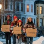 Family in winter clothes smiling while moving boxes with Dream Moving packers in a snowy Astoria neighborhood, illustrating moving with kids during winter break.